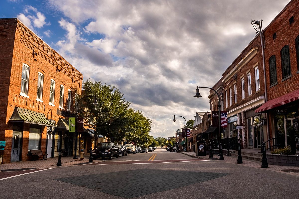 Two-story brick storefronts with awnings lining White Street in downtown Wake Forest, with parked cars, street lamps and a cloudy sky