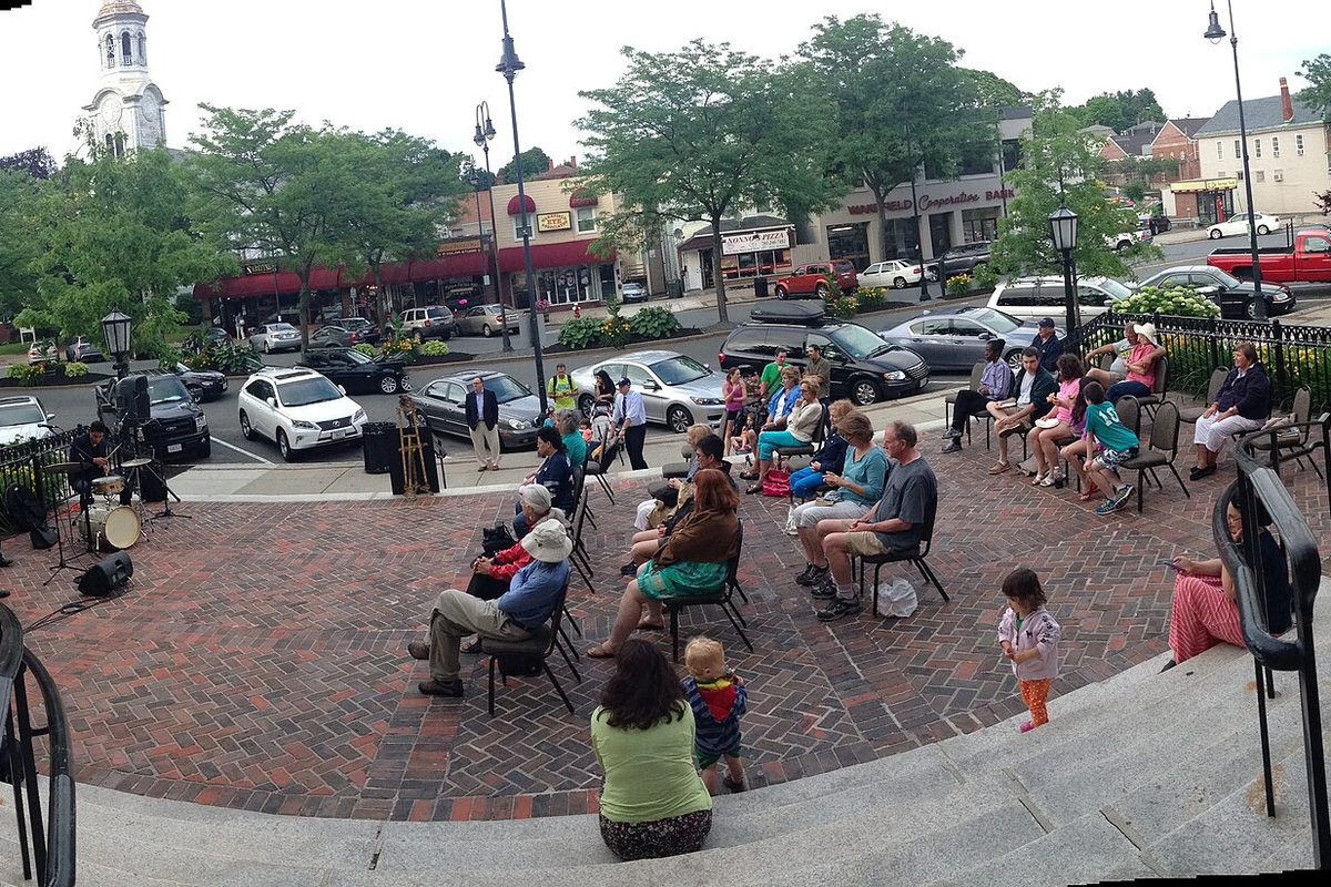People in folding chairs at a jazz concert on the brick plaza of the Beebe Memorial Library, with Main Street, parked cars and a white steeple behind