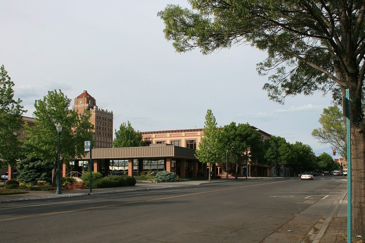 A tree-lined street in downtown Walla Walla with a low flat-roofed building in front of the Marcus Whitman Hotel tower