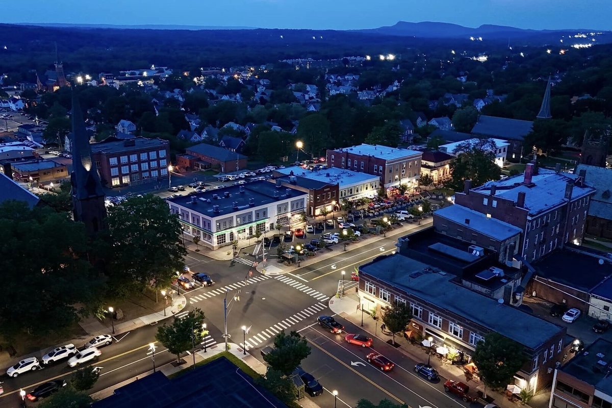 Aerial view of downtown Wallingford at dusk, with lit brick storefronts, a busy intersection with crosswalks, church steeples and wooded hills on the horizon