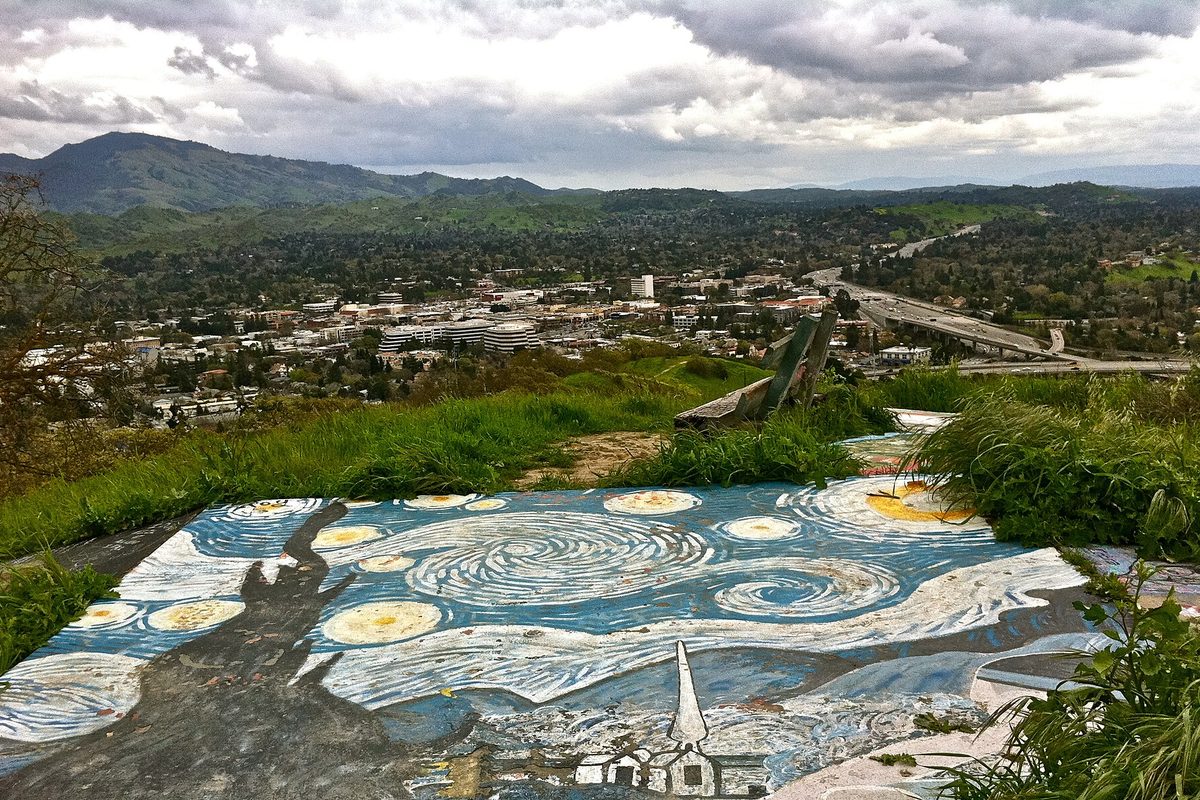 A Starry Night style painting on a concrete slab on a grassy hilltop in Acalanes Open Space, looking out over downtown Walnut Creek and green hills under storm clouds