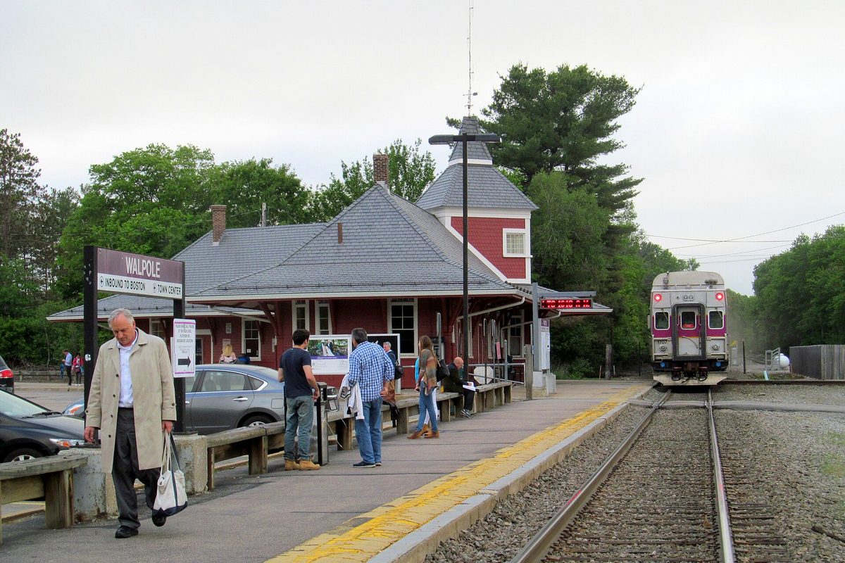 An MBTA commuter train beside the platform at Walpole station, a red depot with a gray roof and cupola, as passengers wait by the station sign