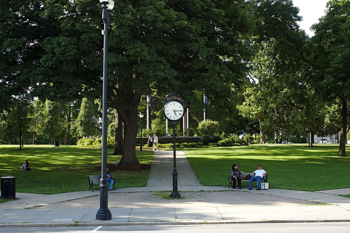 Waltham's Central Square common in summer, a black post clock marked Waltham on a brick walk, shade trees and people sitting on benches