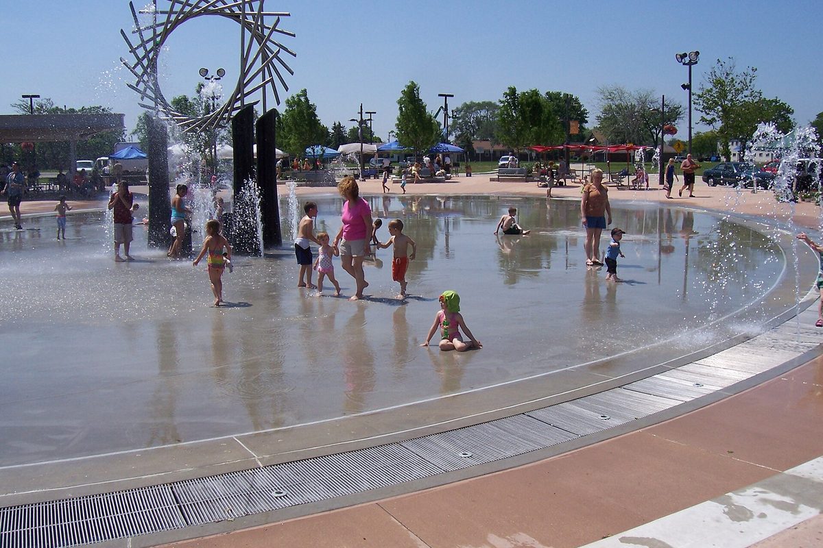 Children playing in a splash pad fountain beside a tall arched metal sculpture at a park in Warren on a sunny day