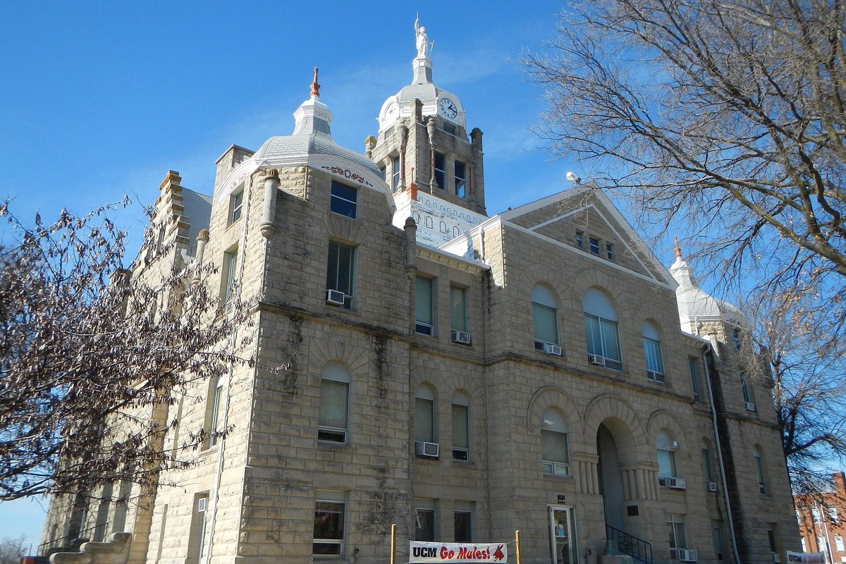 The tan stone Johnson County Courthouse in Warrensburg, with its clock tower and a statue on top, bare trees and a UCM Go Mules banner out front