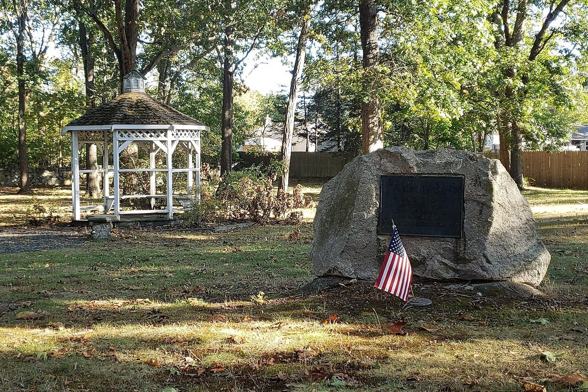 A white gazebo and a large boulder with a dark plaque and a small American flag, under tall trees in Warwick City Park