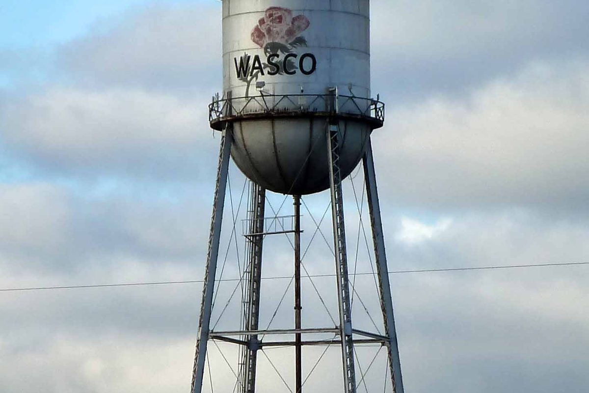 Wasco's silver water tower with the town name and a painted pink rose, standing on steel legs against a cloudy sky