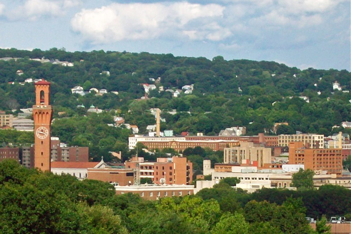 Downtown Waterbury seen from the west, with a tall brick clock tower on the left, red-brick blocks in the middle and houses scattered over a wooded hillside behind