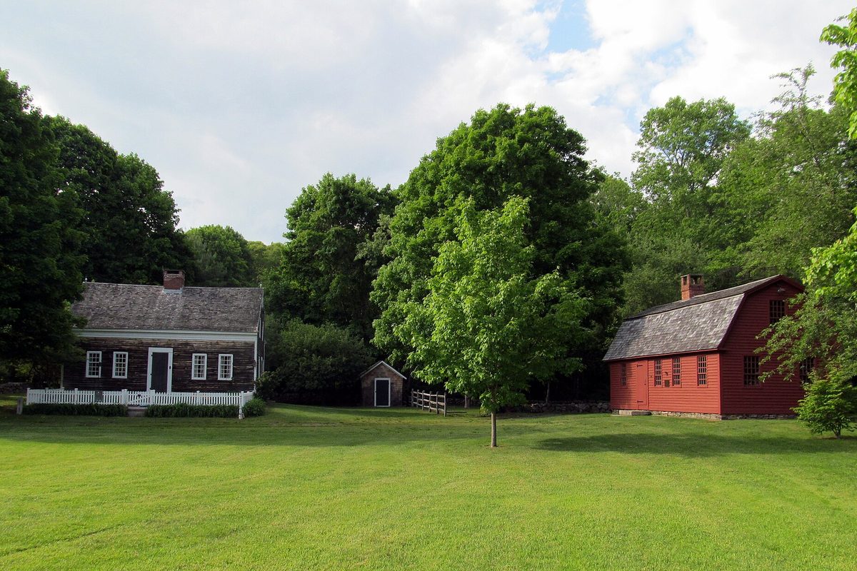 A dark shingled colonial house behind a white picket fence and a small red wooden schoolhouse across a mowed lawn on Jordan Green in Waterford