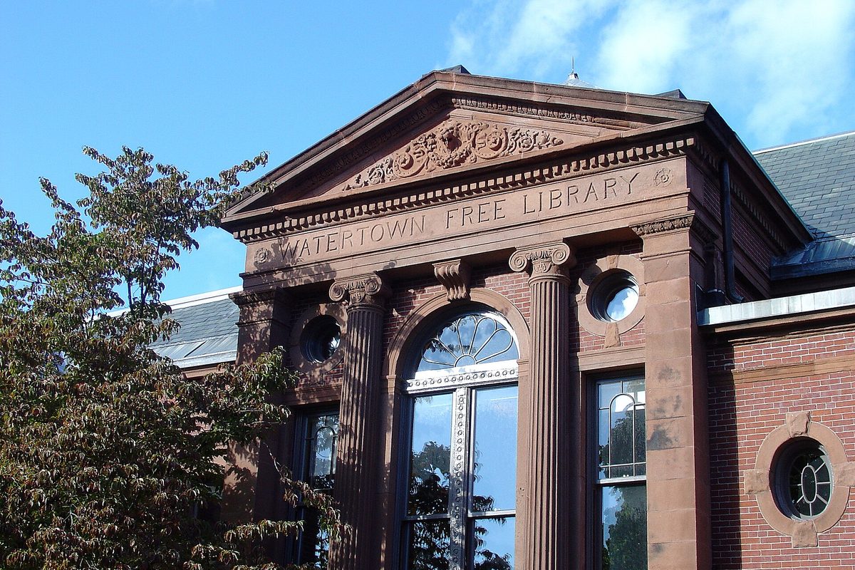 The carved brownstone entrance of the Watertown Free Public Library, with its name across the pediment and an arched window, under blue sky