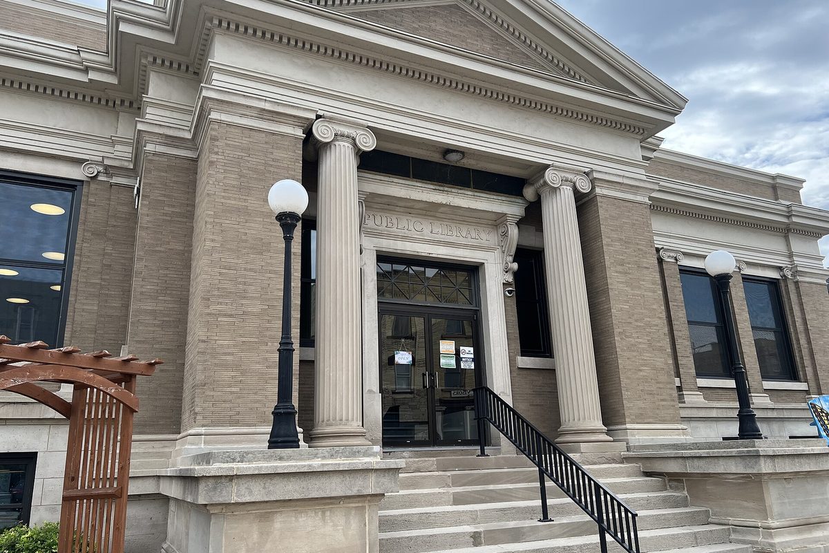 The columned stone entrance of the Watertown Public Library, with Public Library carved above the doors, front steps and two globe lamp posts