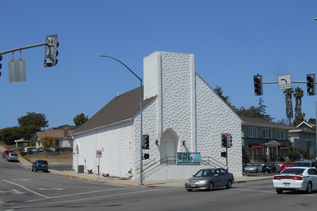 A white windowless church on a Watsonville street corner, with traffic lights overhead and cars passing in front