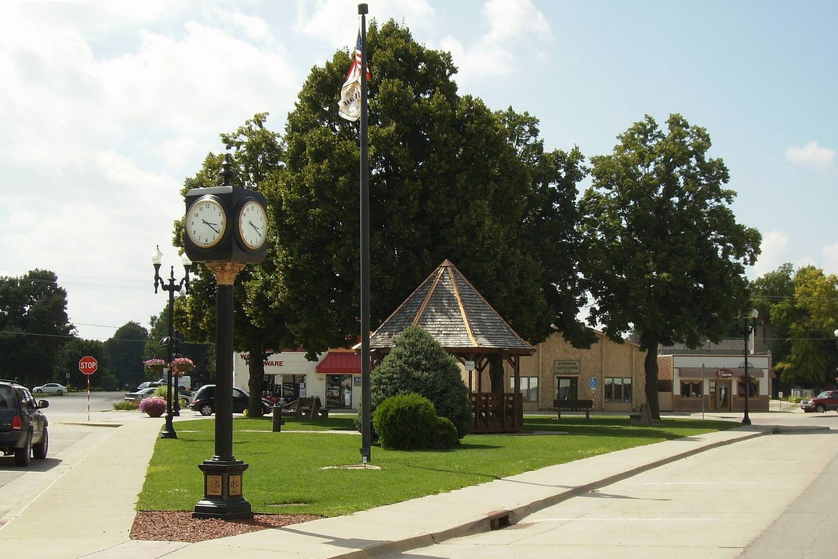 Triangle Park in downtown Waukee, with a two-faced post clock, a flagpole, a wooden gazebo and tall shade trees beside the street
