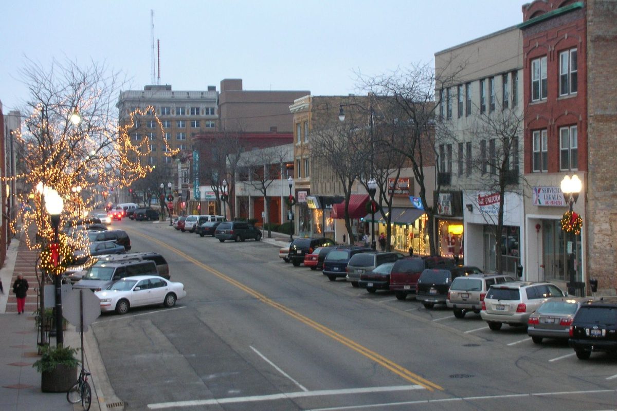 A downtown Waukegan street at dusk, with holiday lights strung through bare trees and cars parked along brick storefronts on both sides