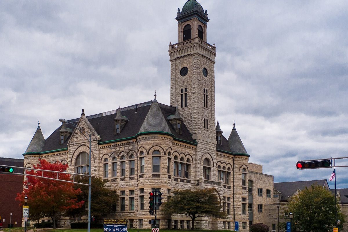 The Old Waukesha County Courthouse, a stone building with turrets and a tall clock tower, under a gray sky with a red-leafed tree at the corner