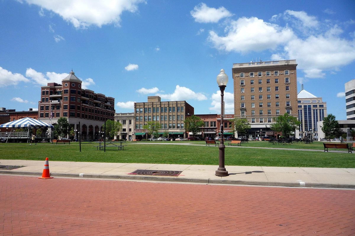 The 400 Block in downtown Wausau, a wide lawn with benches, a lamp post and a tent beside a brick walk, with tall brick buildings behind it