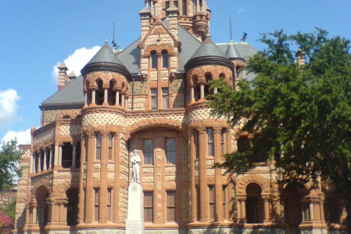 The pink and red stone Ellis County Courthouse in Waxahachie, with turrets, arched windows and a statue on a pillar in front, beside a leafy tree