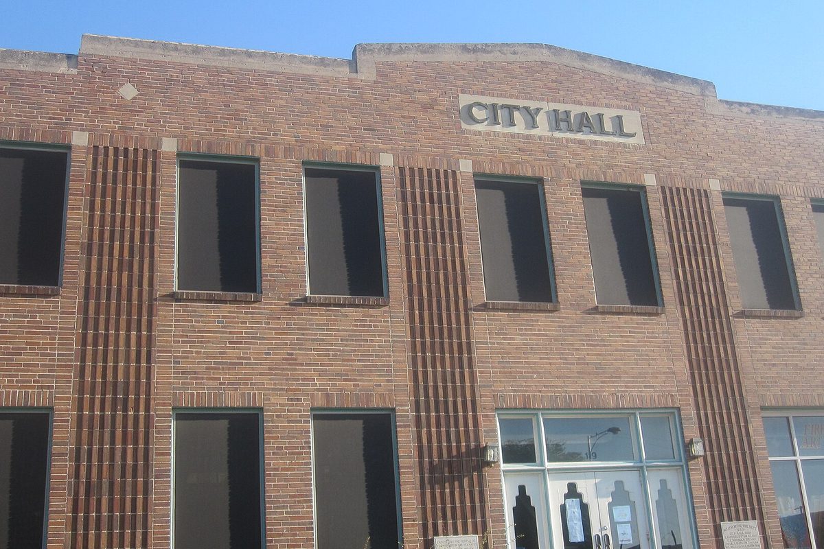 The two-story tan brick front of Weatherford City Hall, with raised CITY HALL lettering, vertical brick piers between tall windows and a glass entry door