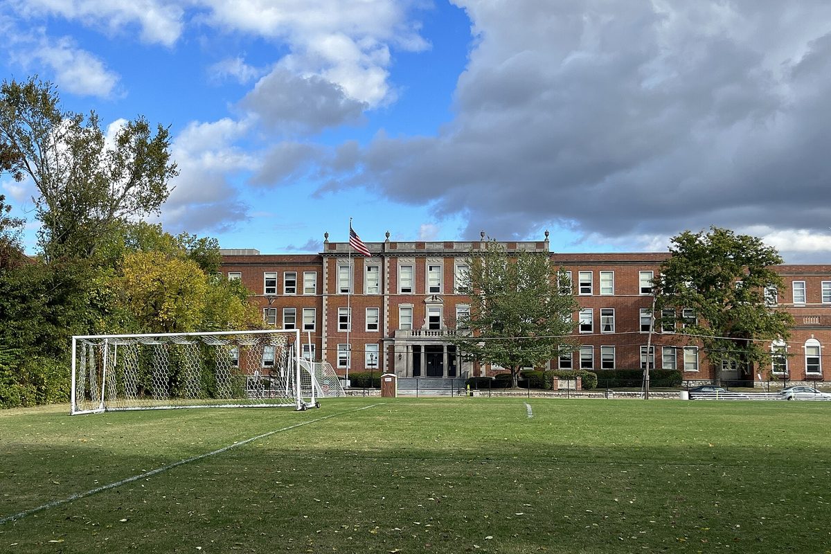 The senior entrance of Webster Groves High School, a long red-brick building with a flag, seen across a soccer field and goal
