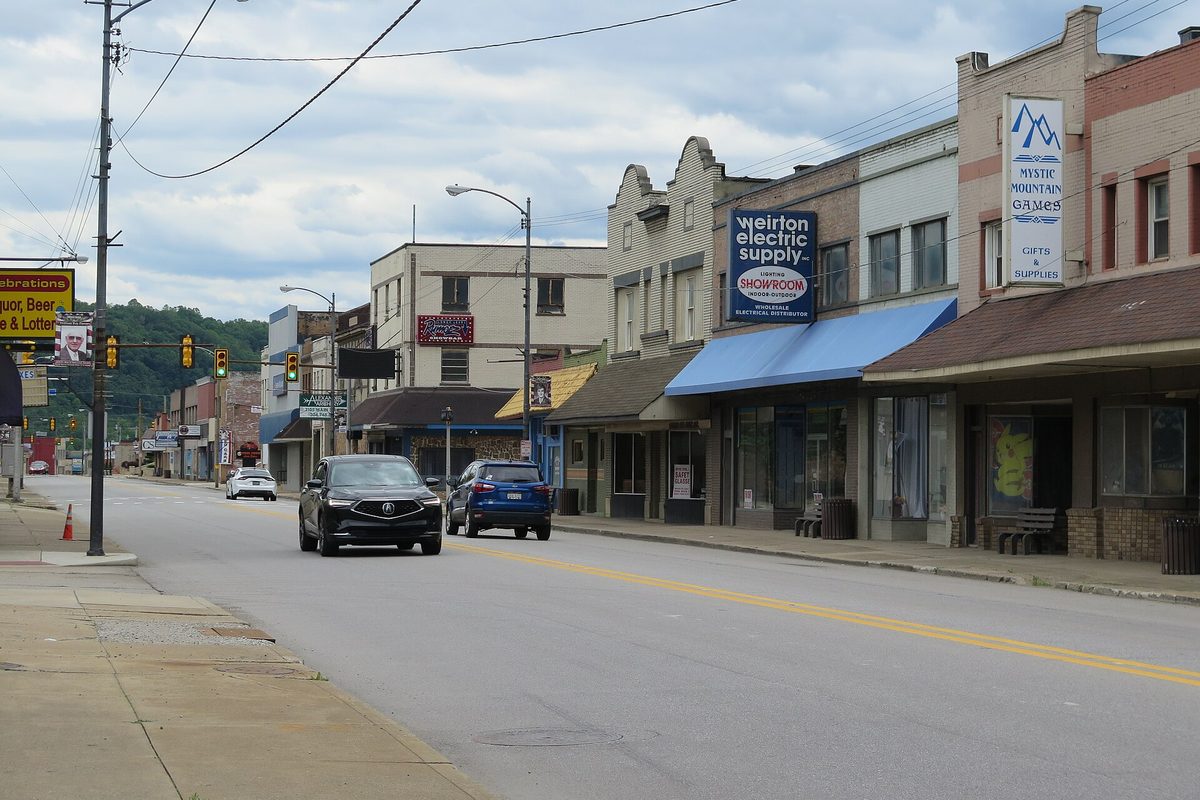 Looking south down Main Street in downtown Weirton, with low brick storefronts, a Weirton Electric Supply sign and two cars on the road