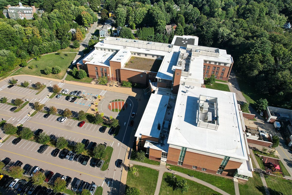 Aerial view of Wellesley High School, a long brick building with white roofs, its parking lot and the trees around it