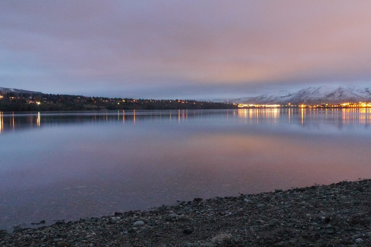 The Columbia River at dusk from a pebble shore in Wenatchee, with the town's lights along the far bank and snow on the hills under a pink sky