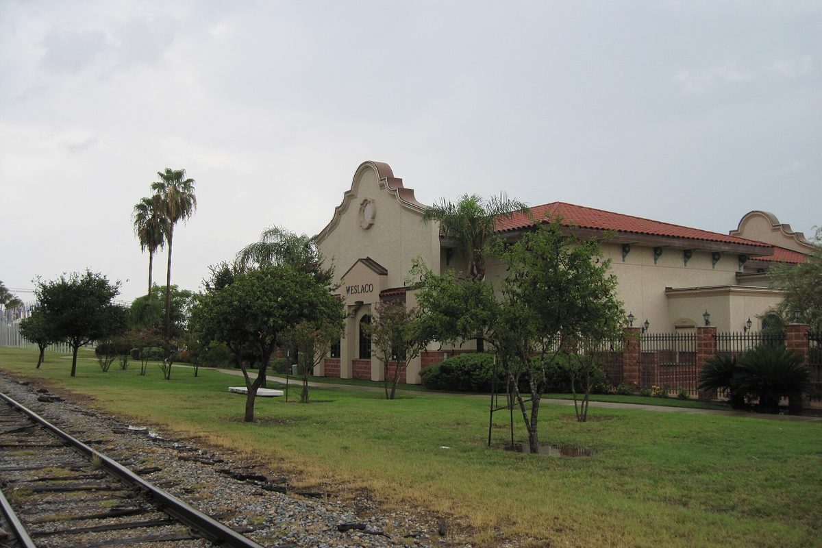 The old Weslaco railroad station, a cream Mission-style building with a curved gable and red tile roof, beside railroad tracks, grass and palm trees under a gray sky