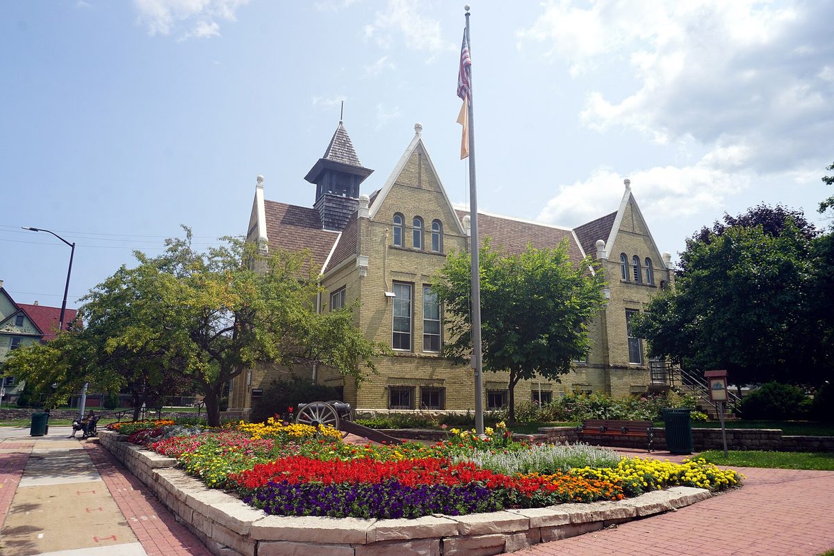 The cream brick Greenfield School, now the West Allis Historical Society Museum, with a bell tower, a flagpole and beds of red, yellow and purple flowers