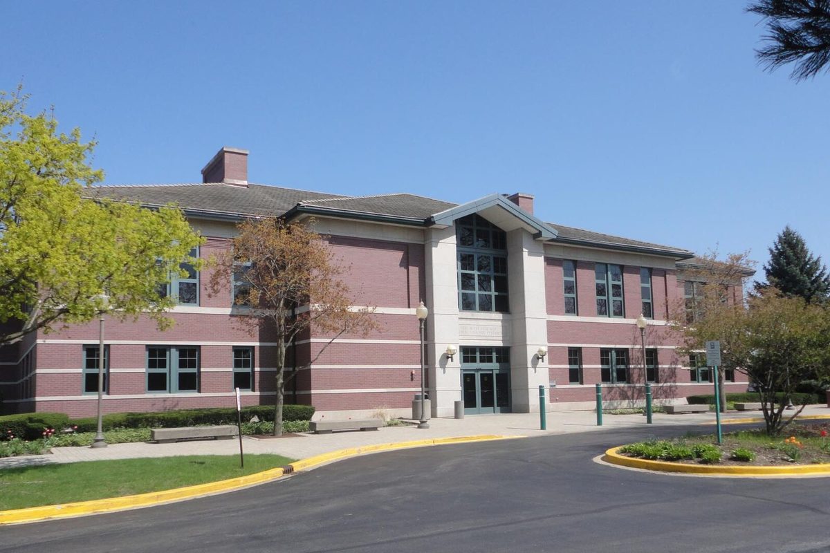 The two-story red brick West Chicago Public Library with a tall glass entrance, a curved drive and spring trees under a clear blue sky