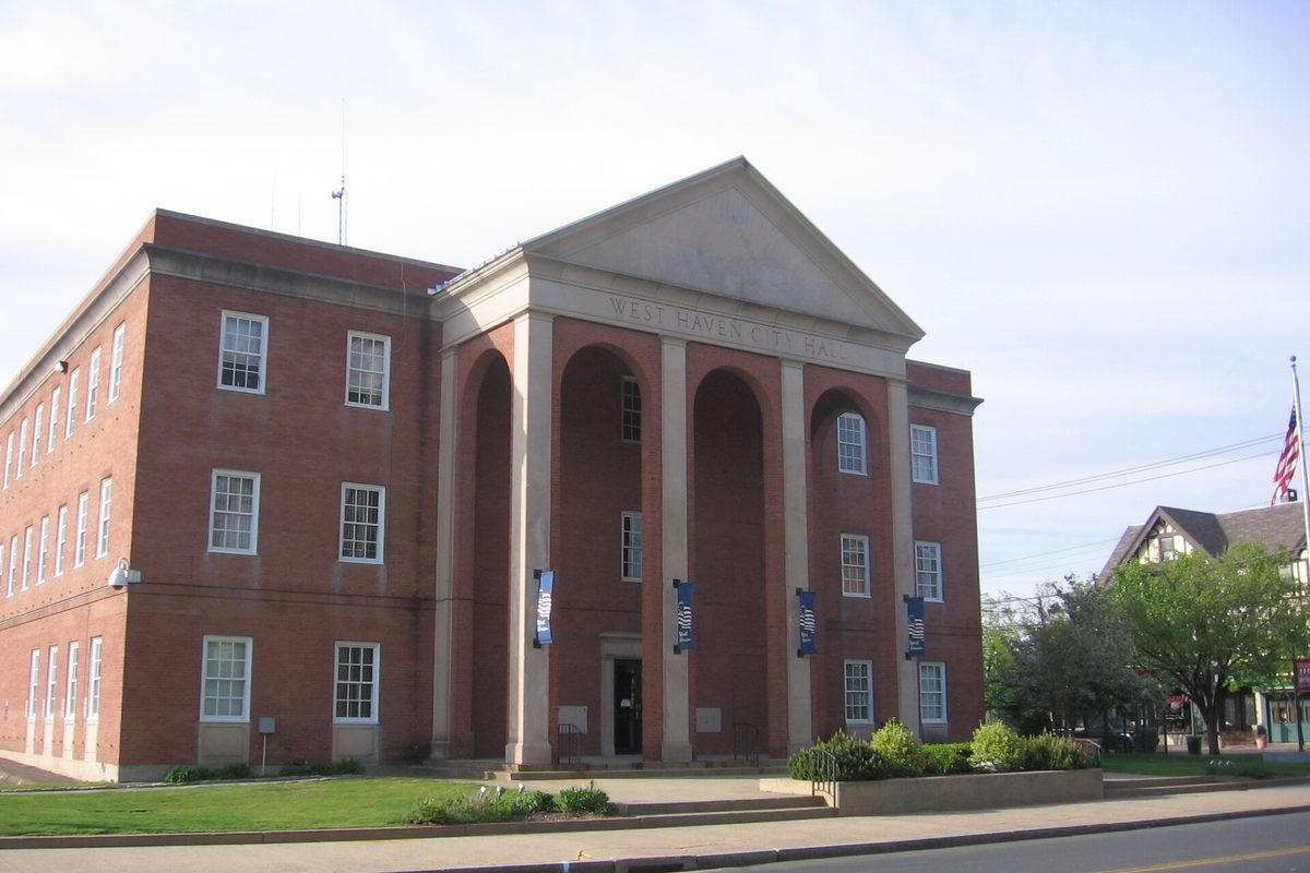 West Haven City Hall, a three-story red brick building with a tall arched portico, blue banners on its columns and an American flag at the right