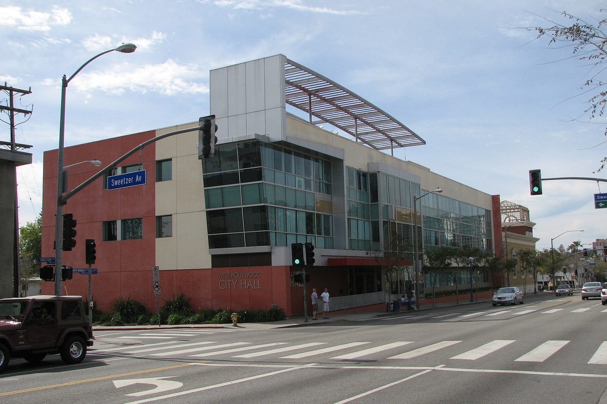 West Hollywood City Hall, a red and cream building with a glass corner and a slatted roof canopy, seen across a crosswalk at the Sweetzer Avenue signal
