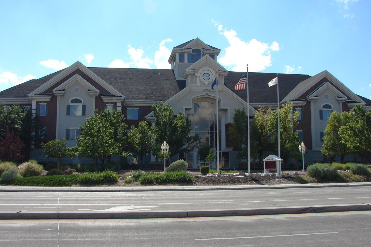 The West Jordan city offices, a gray and brick building with a clock in its central gable, flags out front and a wide road in the foreground