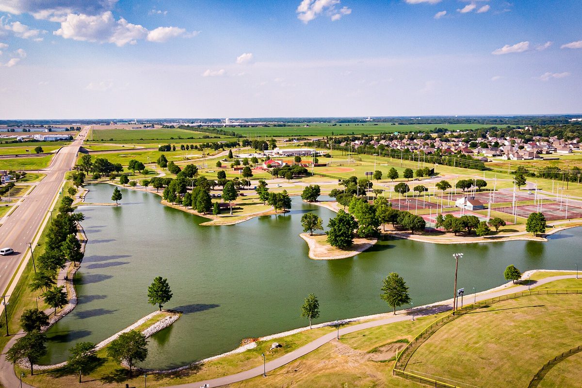 Aerial view of the Tilden Rodgers Sports Complex in West Memphis: a lake with small islands and walking paths, ball fields, and homes, with flat farmland beyond