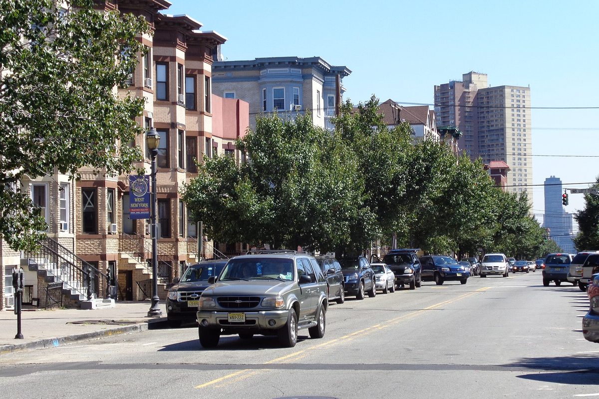 60th Street in West New York looking east, with brownstone rowhouses on the left, a line of parked cars, street trees and high-rise towers in the distance