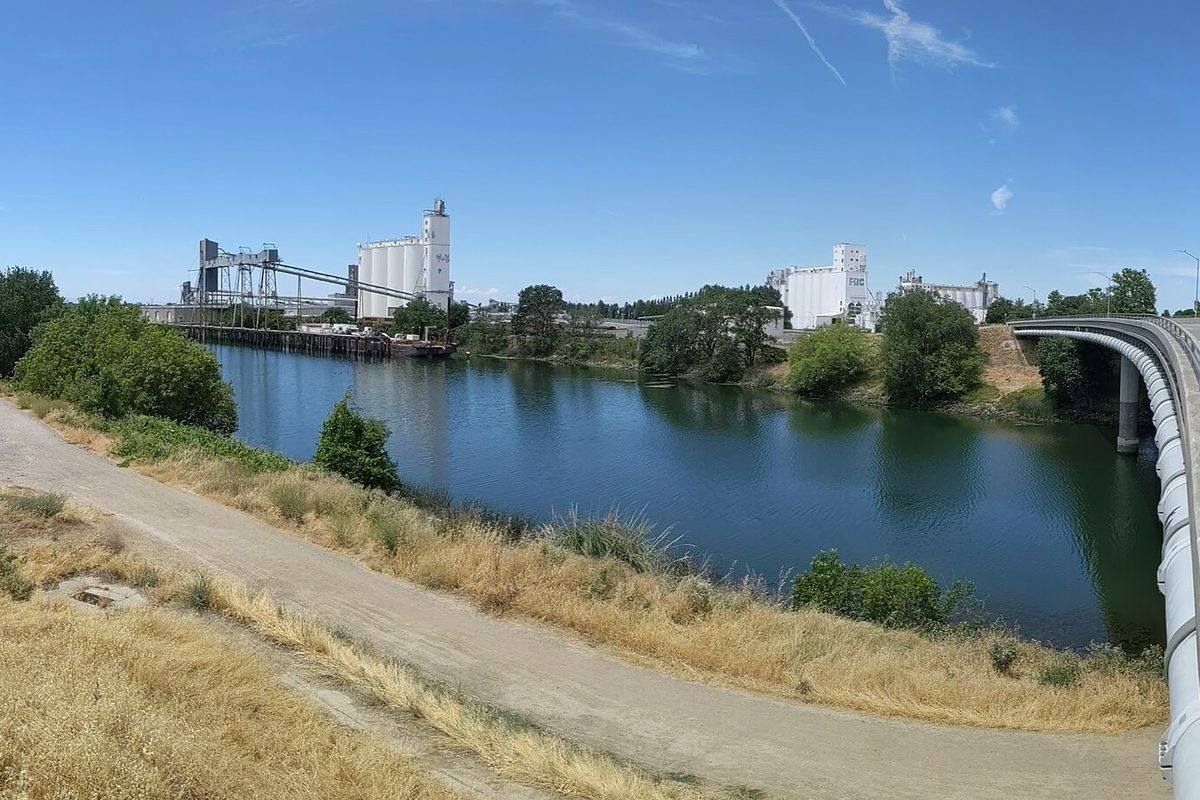 White grain silos and a loading tower at the Port of West Sacramento across the ship channel, seen from a dry-grass path beside a bridge