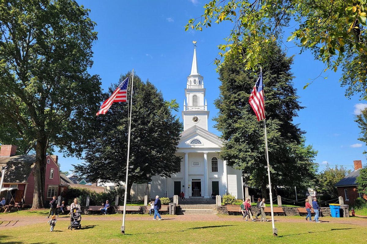 The white Union Meeting House with its tall steeple on Storrowton Green at the Big E, two American flags flying and people walking on the lawn