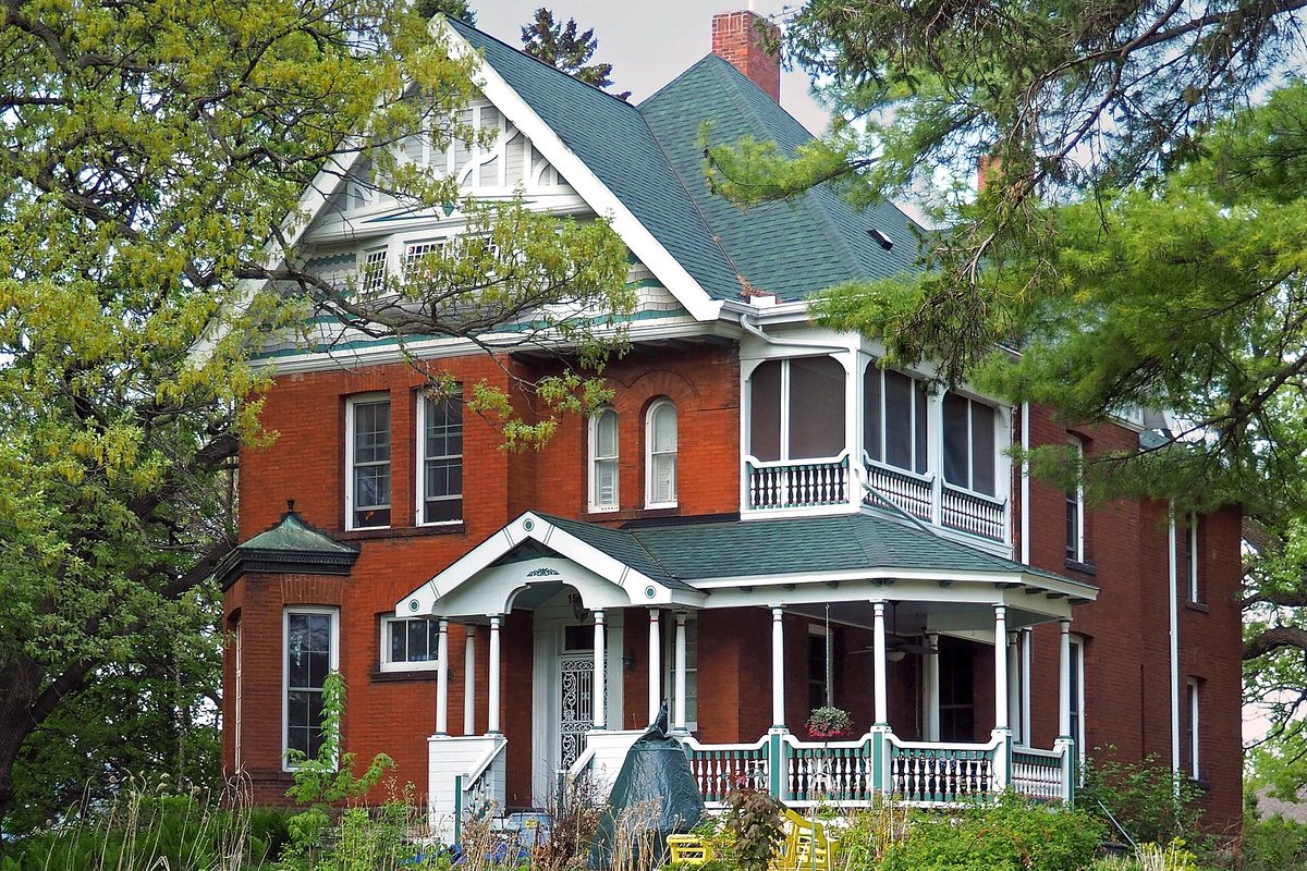 The red-brick George W. Wentworth House in West St. Paul, with a green shingled roof, a white wraparound porch and a turret, half hidden by spring trees