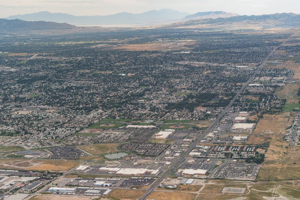 Aerial view looking southeast over West Valley City's grid of homes and warehouses, with a long straight road on the right and mountains on the horizon