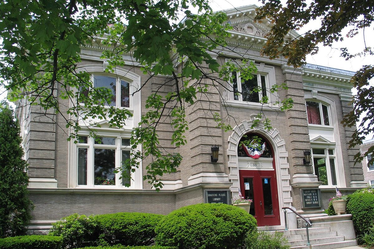 The stone Westborough Public Library with arched windows and red double doors, half hidden behind leafy branches and trimmed hedges