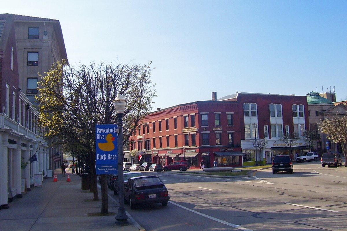 Broad Street in downtown Westerly, with red brick commercial buildings, parked cars and a lamp post sign for the Pawcatuck River Duck Race