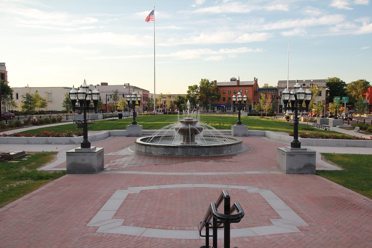 Park Square in Westfield: a fountain ringed by red brick paving and black lamp posts, with a tall flagpole and brick storefronts beyond the green