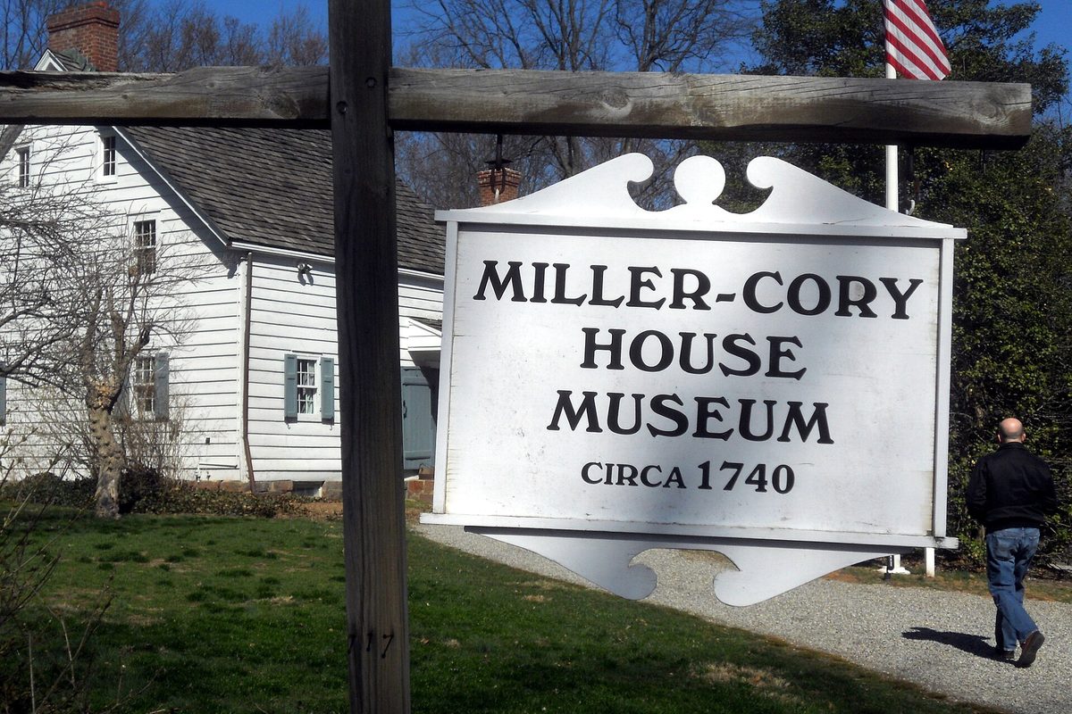 A white hanging sign for the Miller-Cory House Museum on a wooden post in front of a white clapboard house, with a man walking up the gravel path
