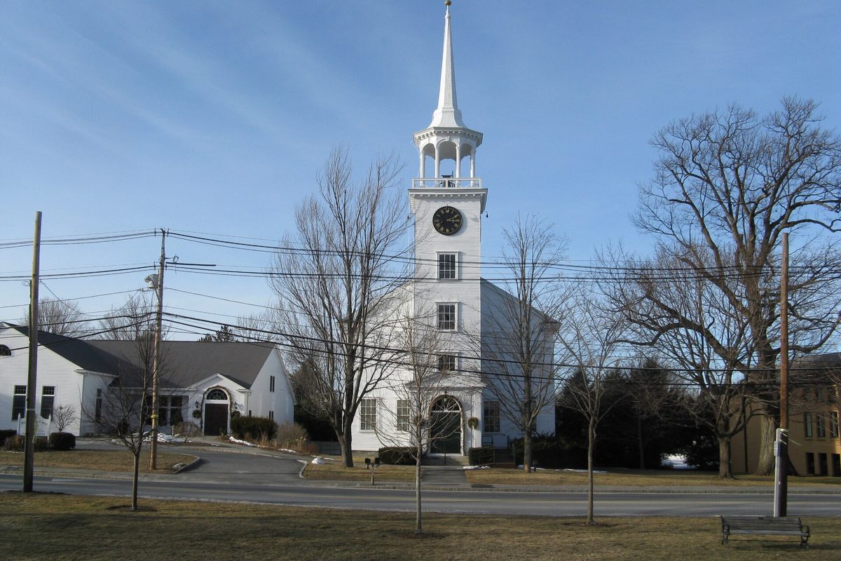 The white First Parish church in Westford Center, with a clock and an open belfry below its spire, behind bare winter trees