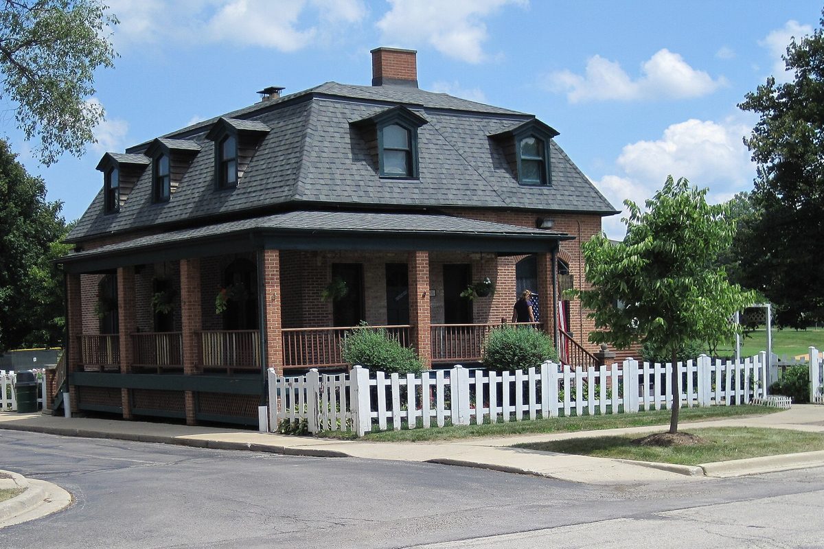 The two-story brick Gregg Museum house in Westmont, with a gray mansard roof, dormer windows, a wide porch and a white picket fence along the sidewalk