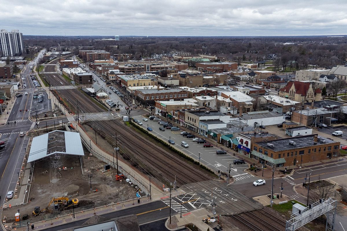Aerial view of downtown Wheaton with the Metra tracks, a station platform under construction, brick storefronts and parking lots under a gray sky