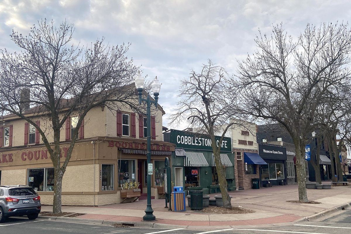 A downtown White Bear Lake street corner with a two-story tan building with maroon shutters, the green Cobblestone Cafe storefront, bare trees and a black lamppost under a cloudy sky