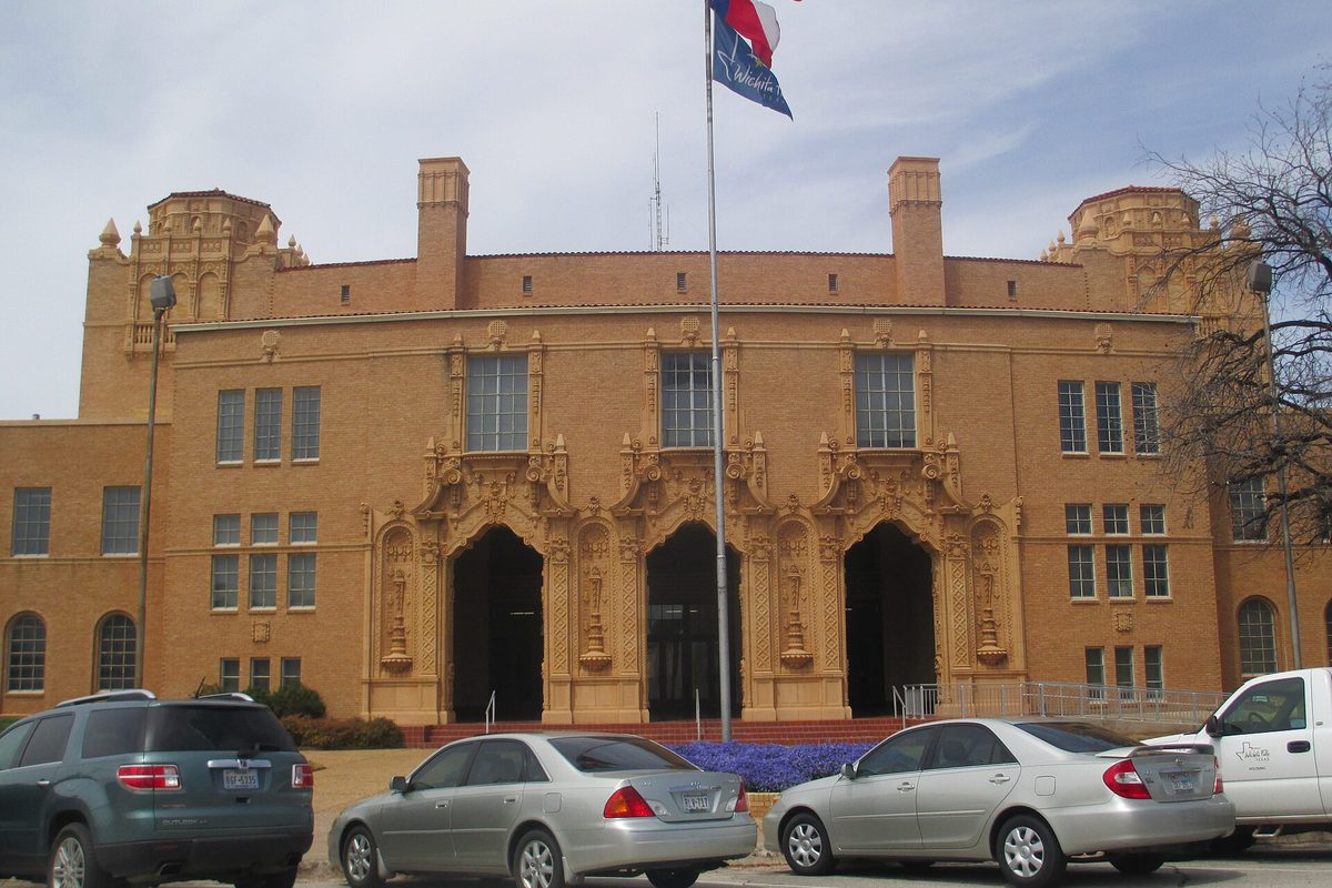 Wichita Falls City Hall in the tan brick Municipal Auditorium, with ornate carved trim around three arched entrances, a Texas flag on a tall pole and cars parked in front