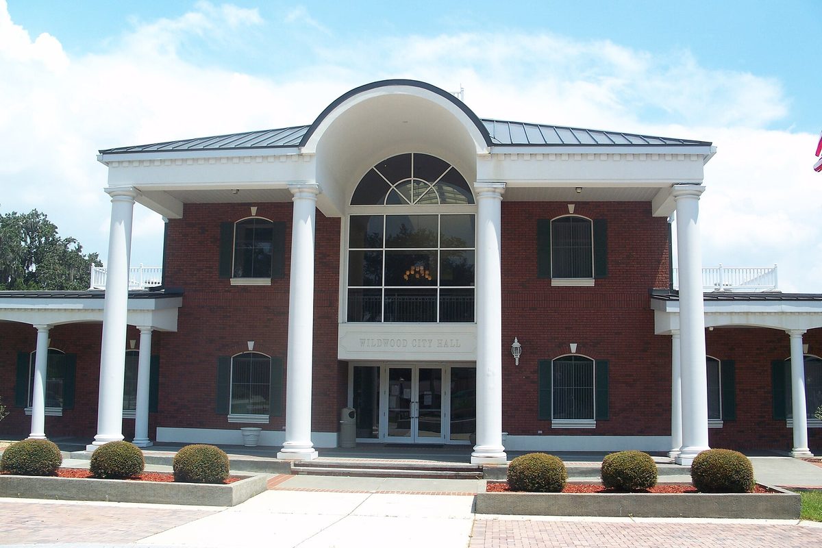 Wildwood City Hall, a two-story red brick building with tall white columns, a white arched entry with a fan window, and trimmed shrubs along the front walk