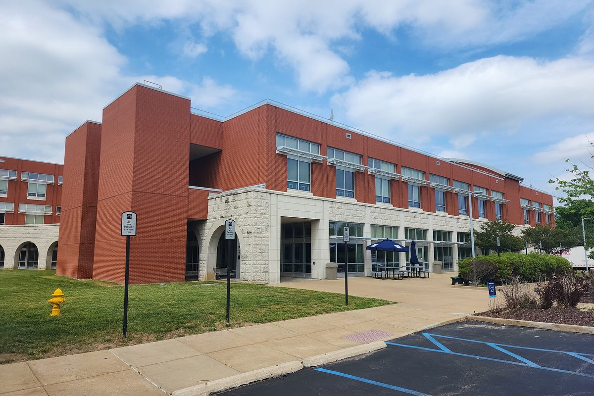 St. Louis Community College at Wildwood, a red brick and pale stone building with a blue patio umbrella out front, under a partly cloudy sky