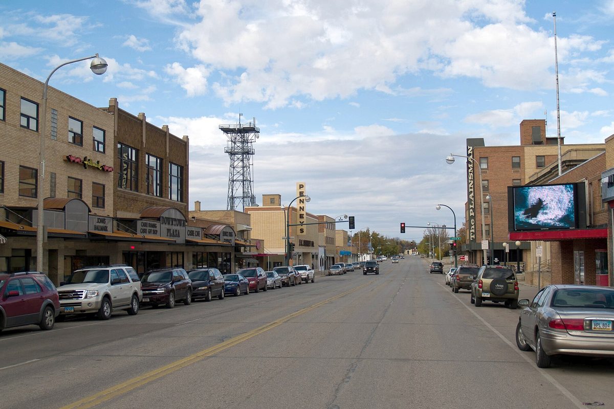 A wide Williston main street with brick storefronts, a theater sign, a steel radio tower and SUVs parked along both curbs